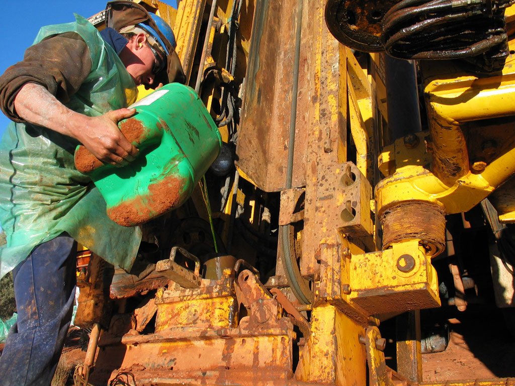 A worker wearing safety gear and a blue hard hat pours lubricant or hydraulic fluid from a green container into a large, yellow industrial drilling machine. The machinery and the worker’s clothing are covered in reddish-brown mud, indicating a rugged working environment. Image credit: LNG Canada Alliance