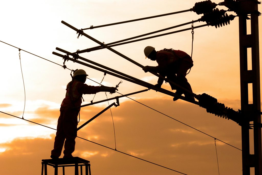 Two utility workers wearing safety gear work on high-voltage power lines at sunset, silhouetted against an orange sky.