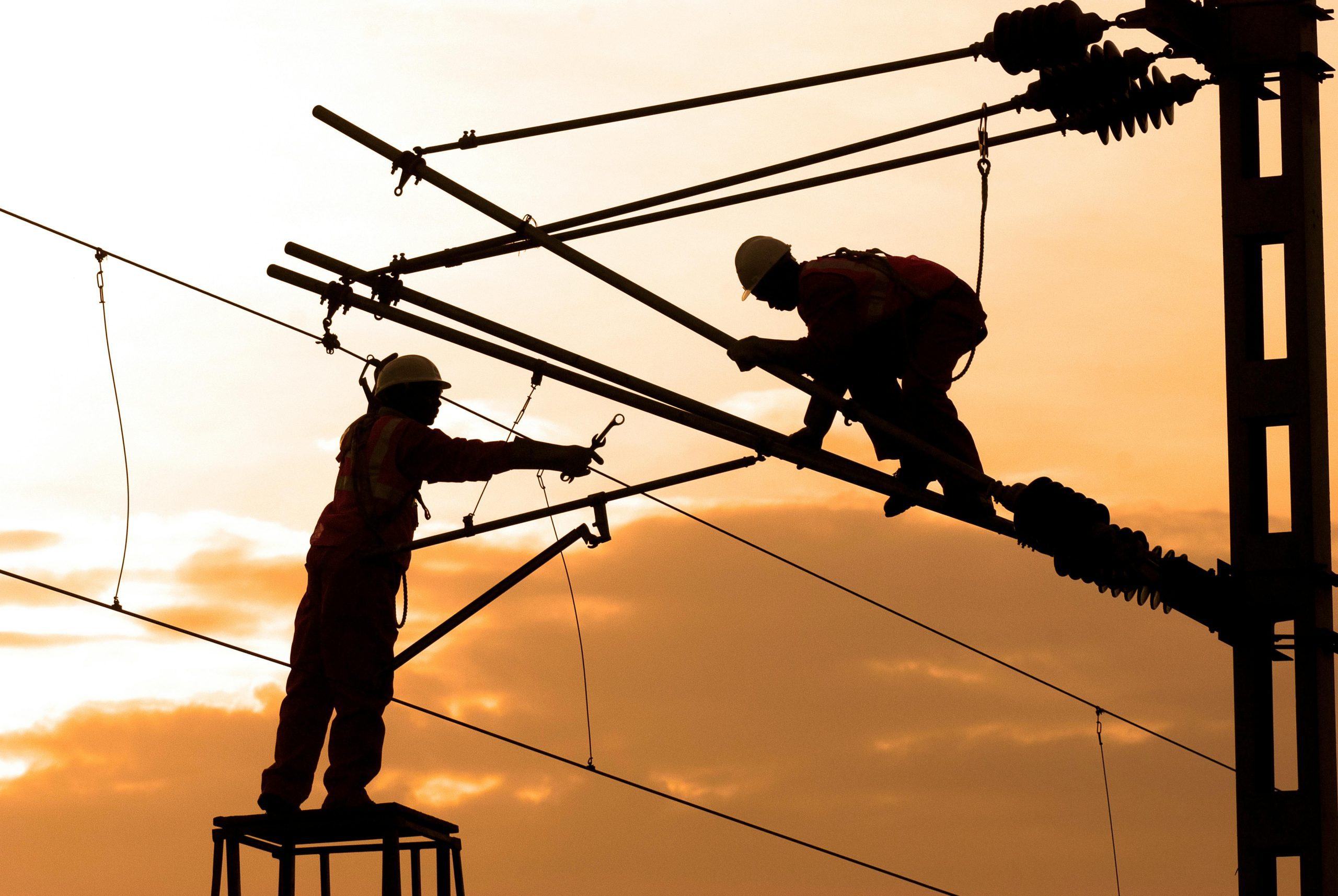 Two utility workers wearing safety gear work on high-voltage power lines at sunset, silhouetted against an orange sky.